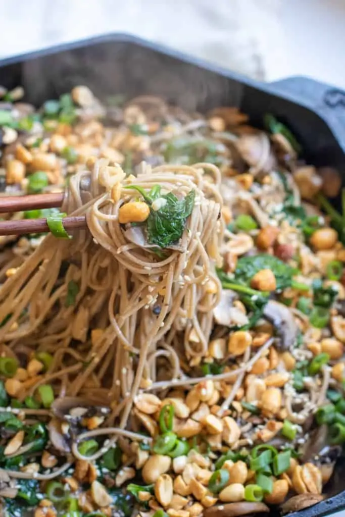 cast iron of noodles with spinach, mushrooms, topped with peanuts and scallions chopsticks holding up noodles.