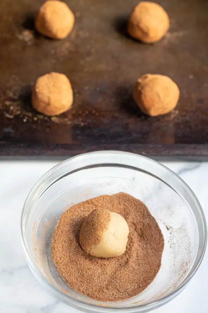 a small dough ball being rolled in a sugar and cinnamon bowl and coated doughs on a baking sheet.