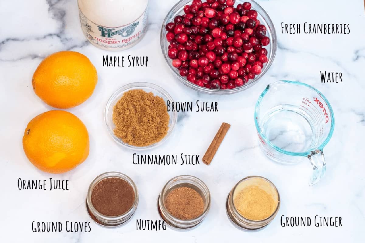 cranberries, oranges, maple syrup, spices, sugar, and cinnamon stick on a counter.