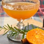 an orange cocktail with a cinnamon and sugar rim on a board with rosemary and halved orange.