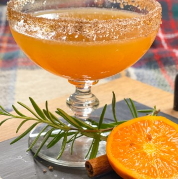 an orange cocktail with a cinnamon and sugar rim on a board with rosemary and halved orange.