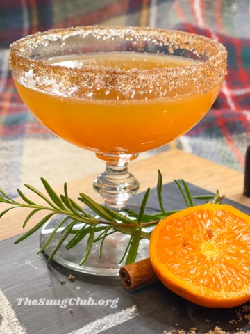 an orange cocktail with a cinnamon and sugar rim on a board with rosemary and halved orange.