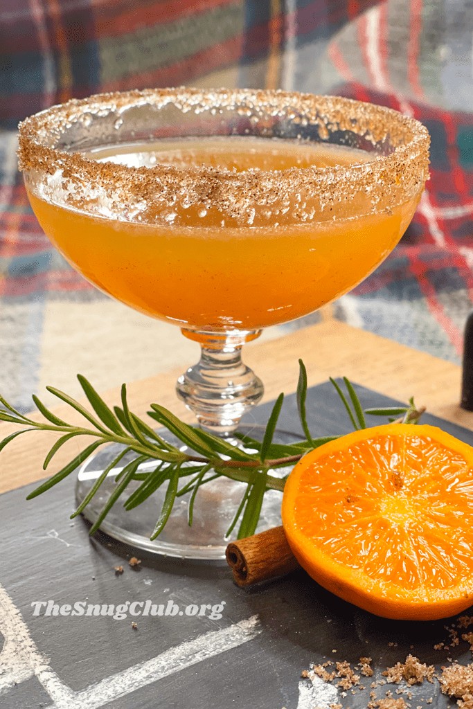 an orange cocktail with a cinnamon and sugar rim on a board with rosemary and halved orange.