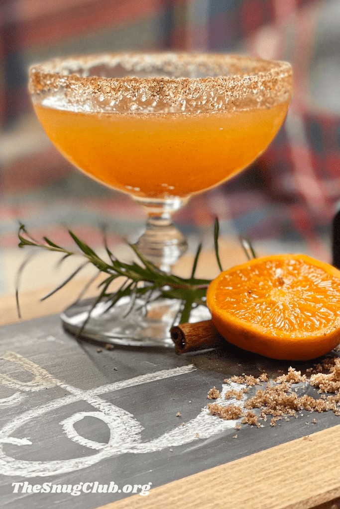 an orange cocktail with a cinnamon and sugar rim on a board with rosemary and halved orange.