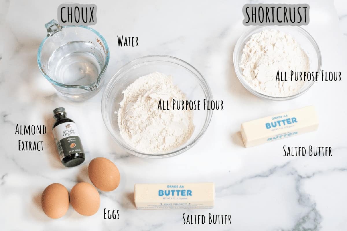 two sticks of butter, two bowls of flour, eggs, extract, and water on a counter.
