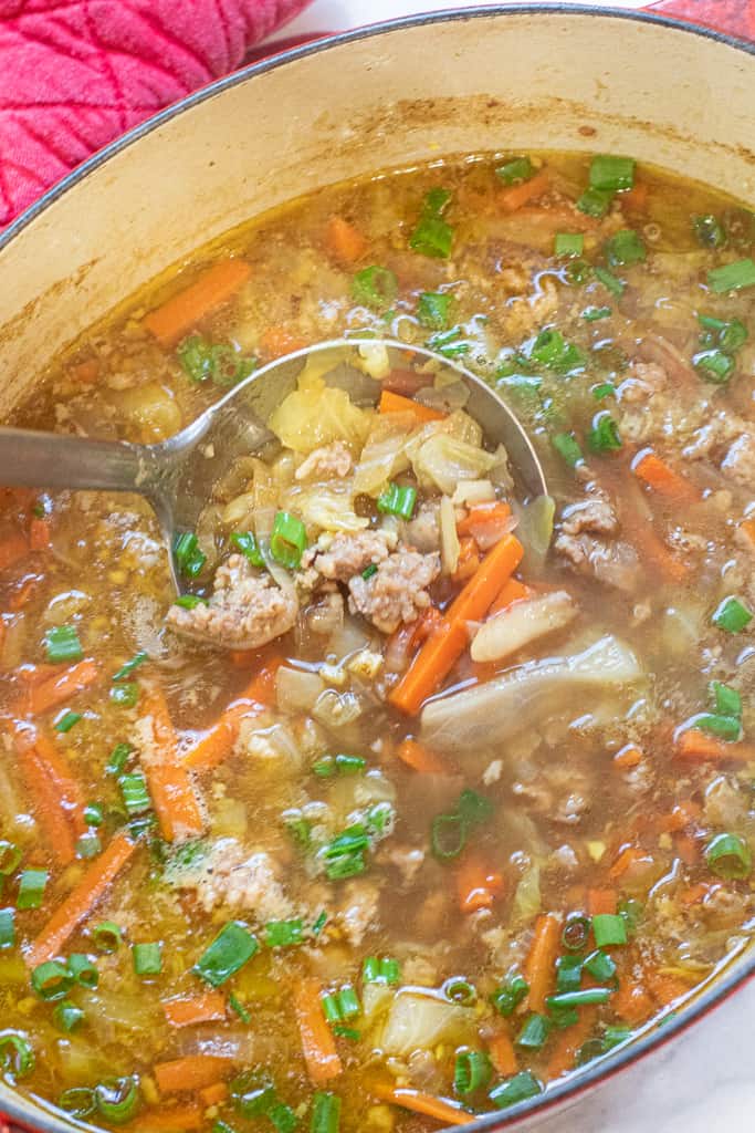 a ladle of soup in dutch oven with cabbage, carrots, and pork.