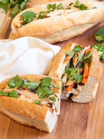 a halved banh mi and a full banh mi on a cutting board with sliced jalapeno and cilantro.