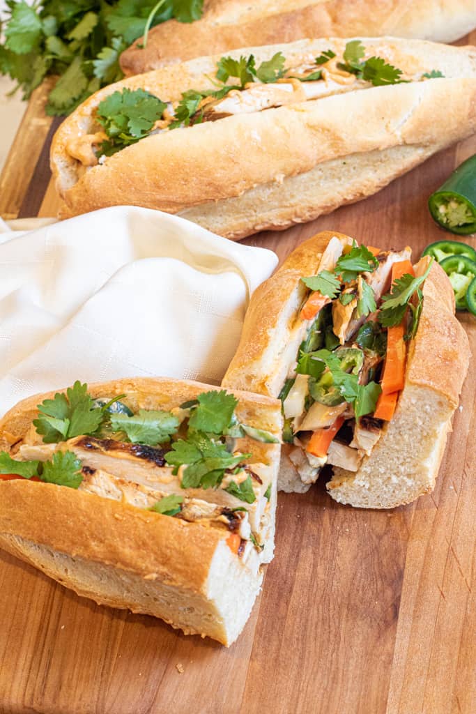 a halved banh mi and a full banh mi on a cutting board with sliced jalapeno and cilantro.