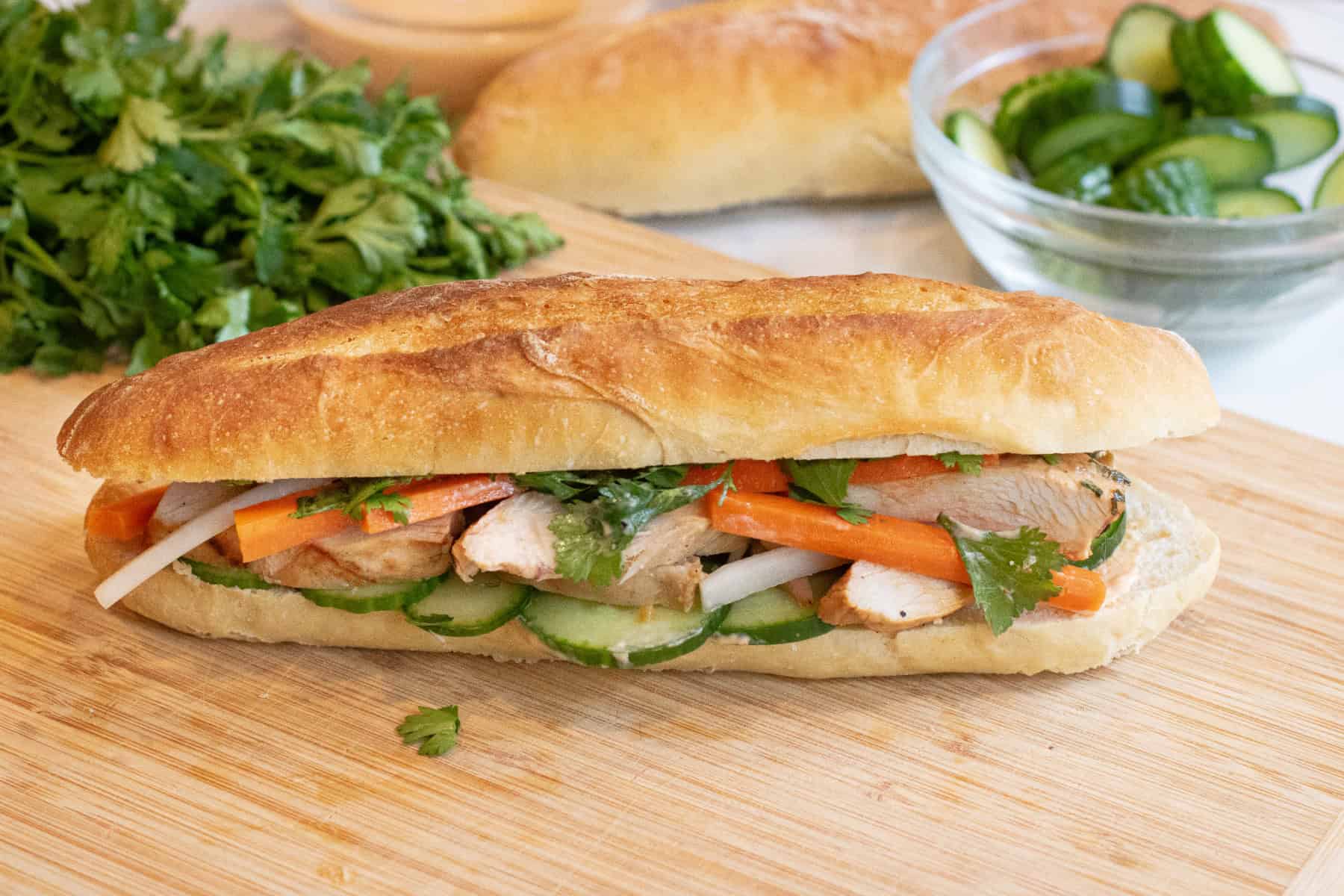 a chicken banh mi sandwich with cucumbers and carrots and cilantro on a cutting board.
