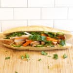 a chicken banh mi sandwich with cucumbers and carrots and cilantro on a cutting board.