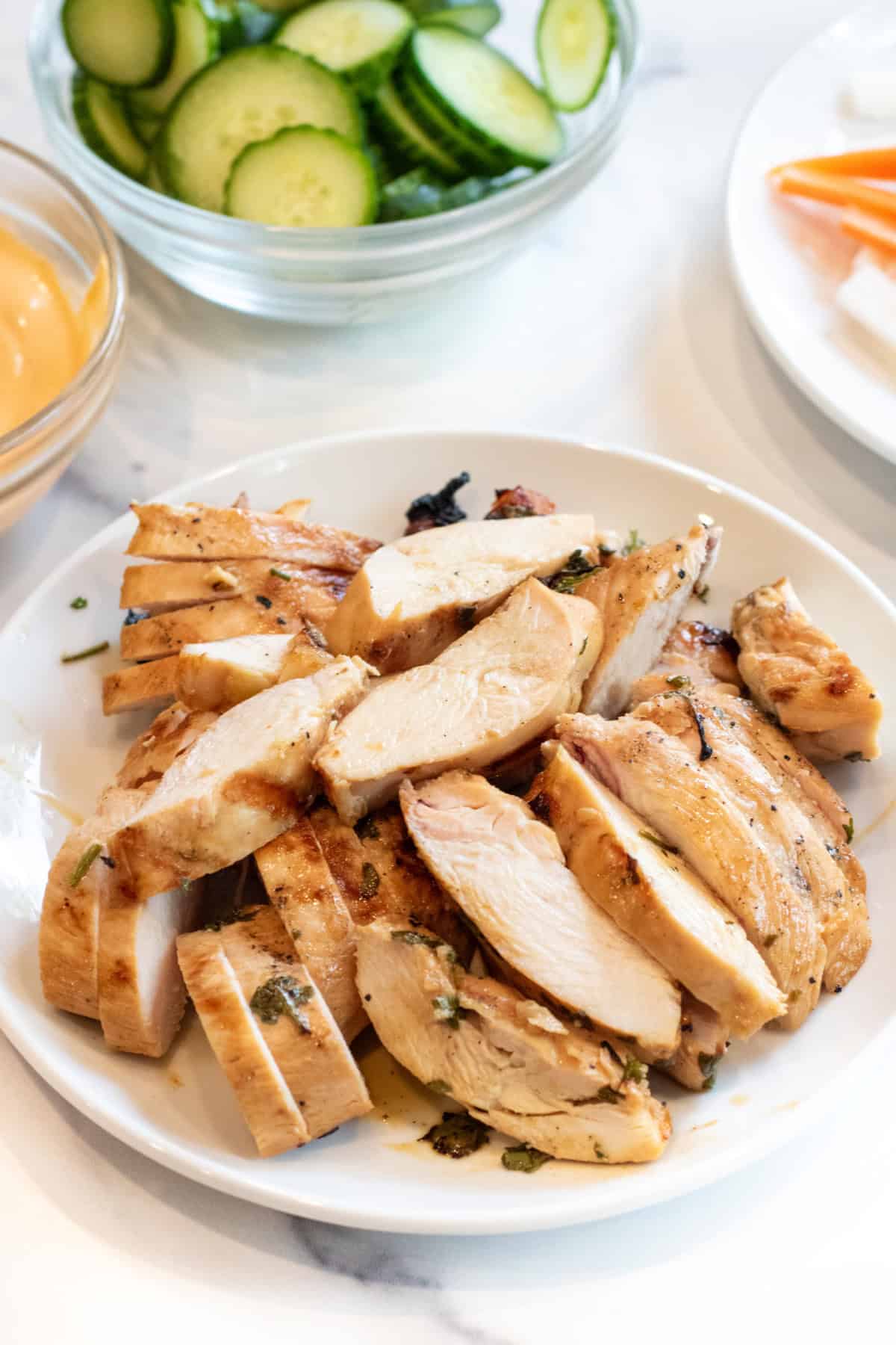 a plate of sliced grilled chicken in front of bowls of mayo, cucumber, and carrot and daikon.