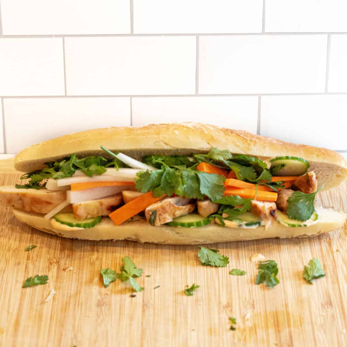 a chicken banh mi sandwich with cucumbers and carrots and cilantro on a cutting board.