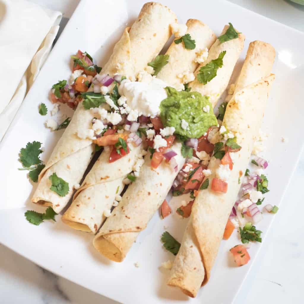 baked flautas on a plate topped with pico, guac, and sour cream.