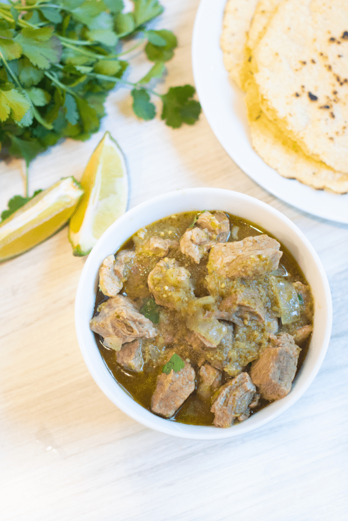 a bowl of pork verde next to limes, tortillas, and cilantro.