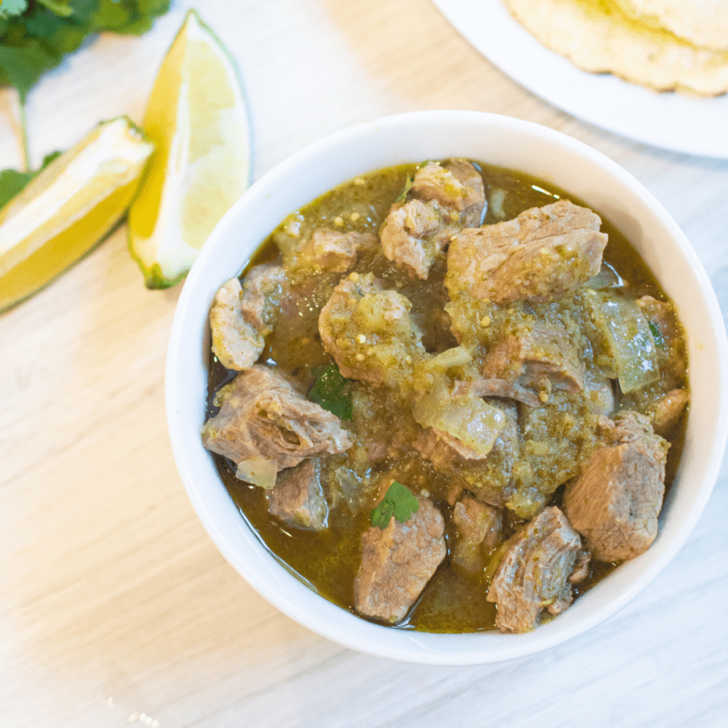 a bowl of pork verde next to limes, tortillas, and cilantro.