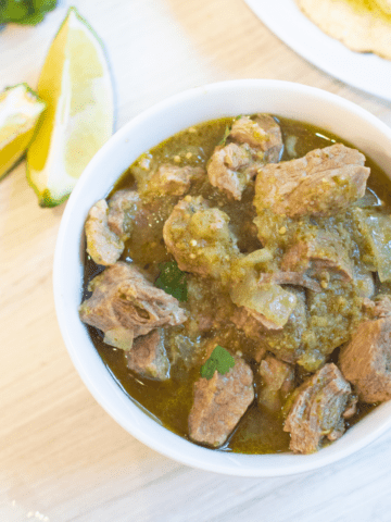 a bowl of pork verde next to limes, tortillas, and cilantro.