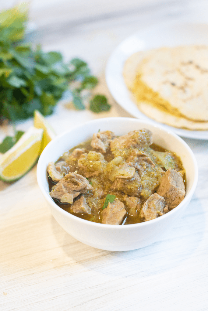 a bowl of pork verde next to limes, tortillas, and cilantro.