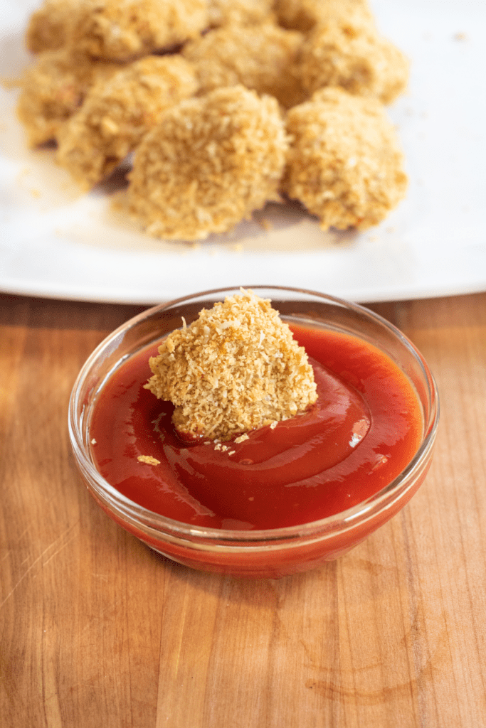 a chicken nugget in a bowl of ketchup next to a plate of chicken nuggets.