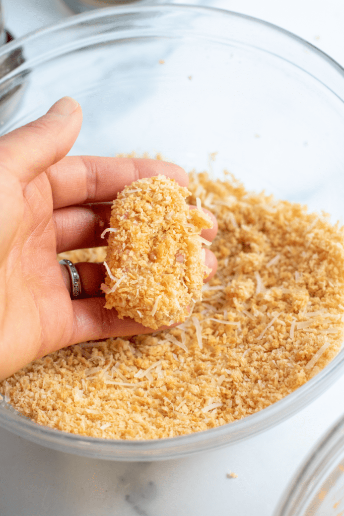 a hand holding a shaped and breaded chicken nugget over a bowl of bread crumbs.