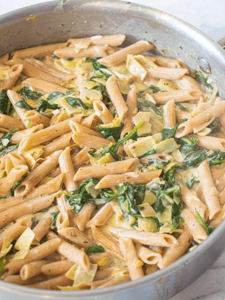 a close up of a pan of penne with spinach and artichokes in a creamy sauce.