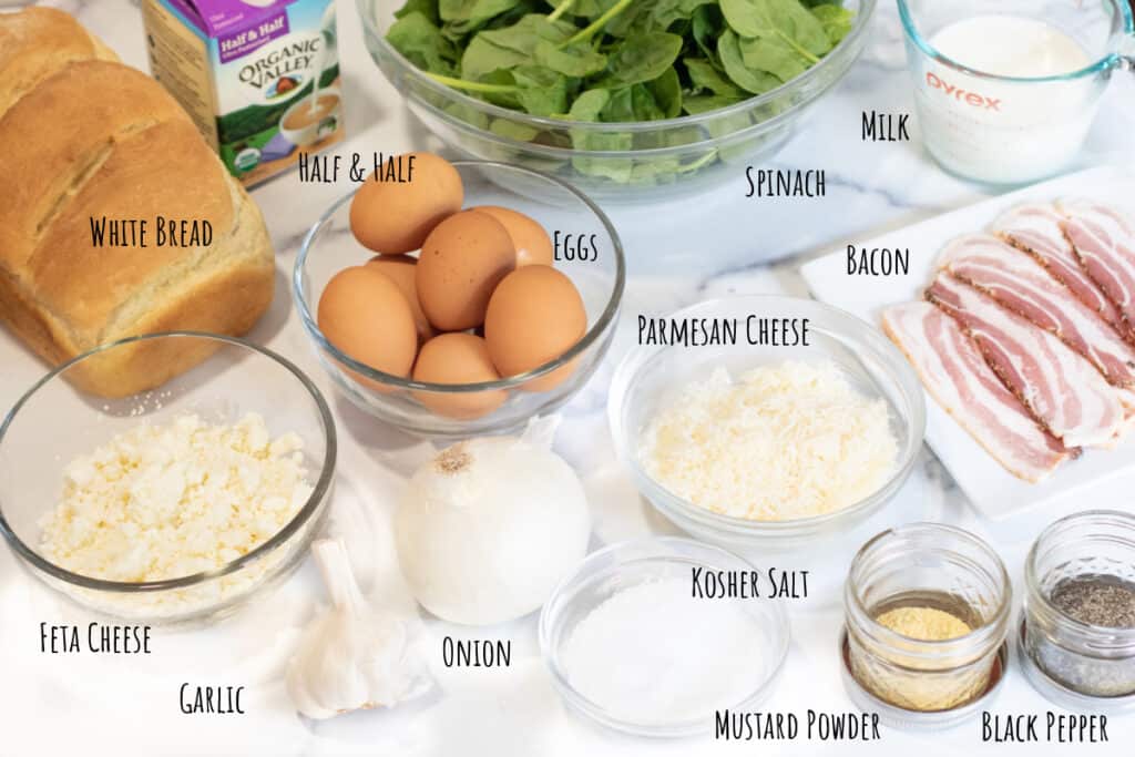 bread, eggs, cheeses, spinach, milk, cream, bacon, spices, onion, and garlic on a counter.