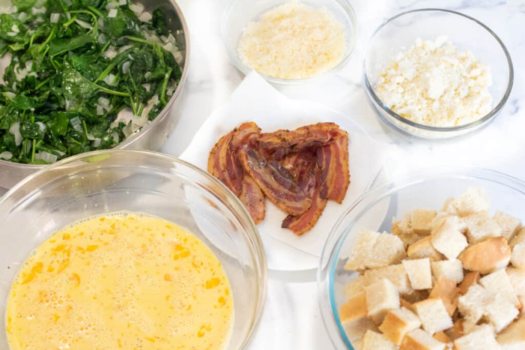 a pan of cooked spinach, whisked eggs, cooked bacon, cheese, and cubed bread.