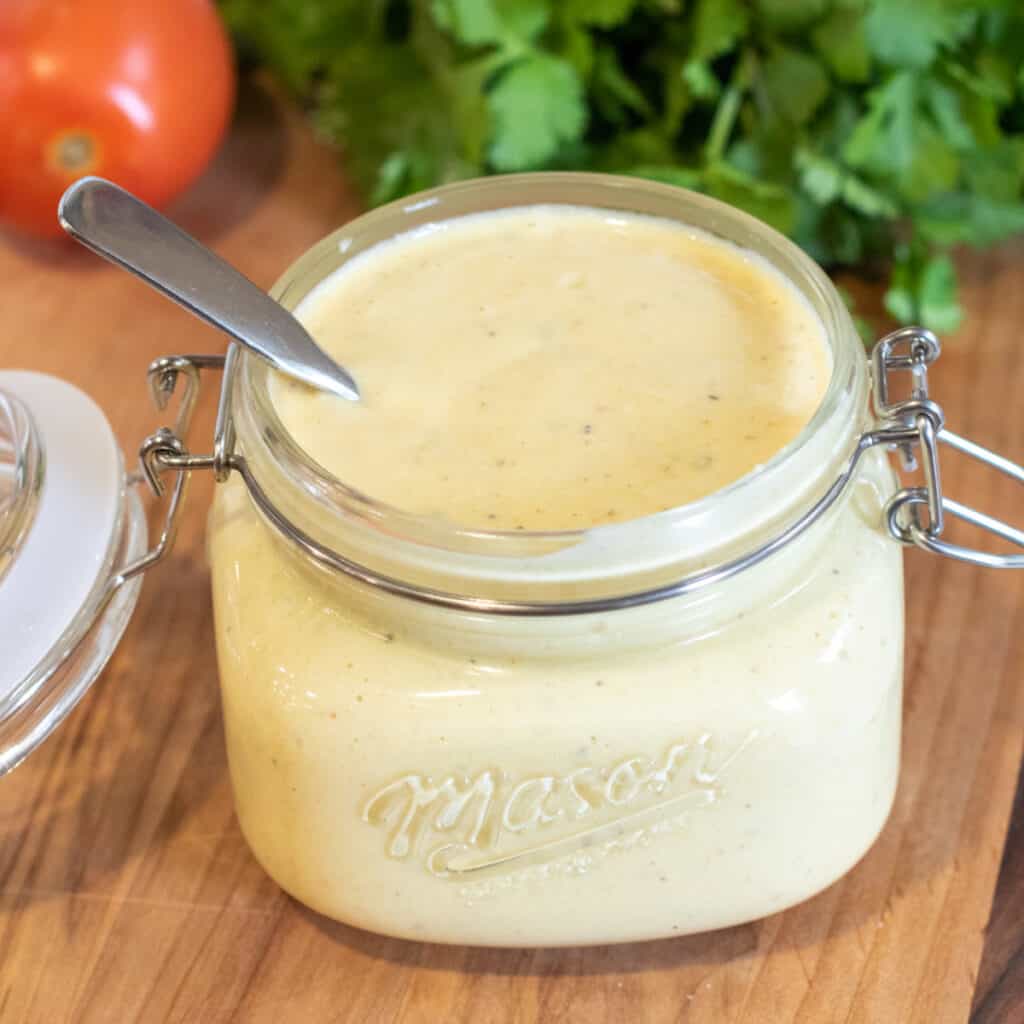 an open jar of sauce with a spoon in it in front of cilantro and tomatoes.