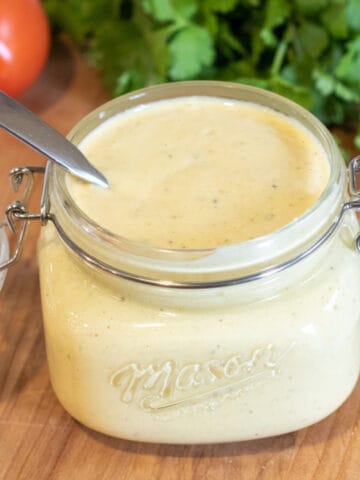 an open jar of sauce with a spoon in it in front of cilantro and tomatoes.