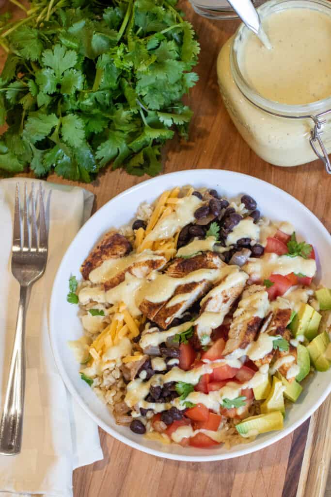 a bowl of chicken, beans, tomatoes, avocado, and sauce next to a jar of sauce and fresh cilantro.