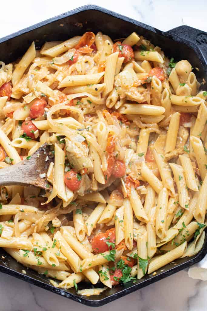 a cast iron of penne pasta with tomatoes and artichokes and parsley with a wooden spoon.