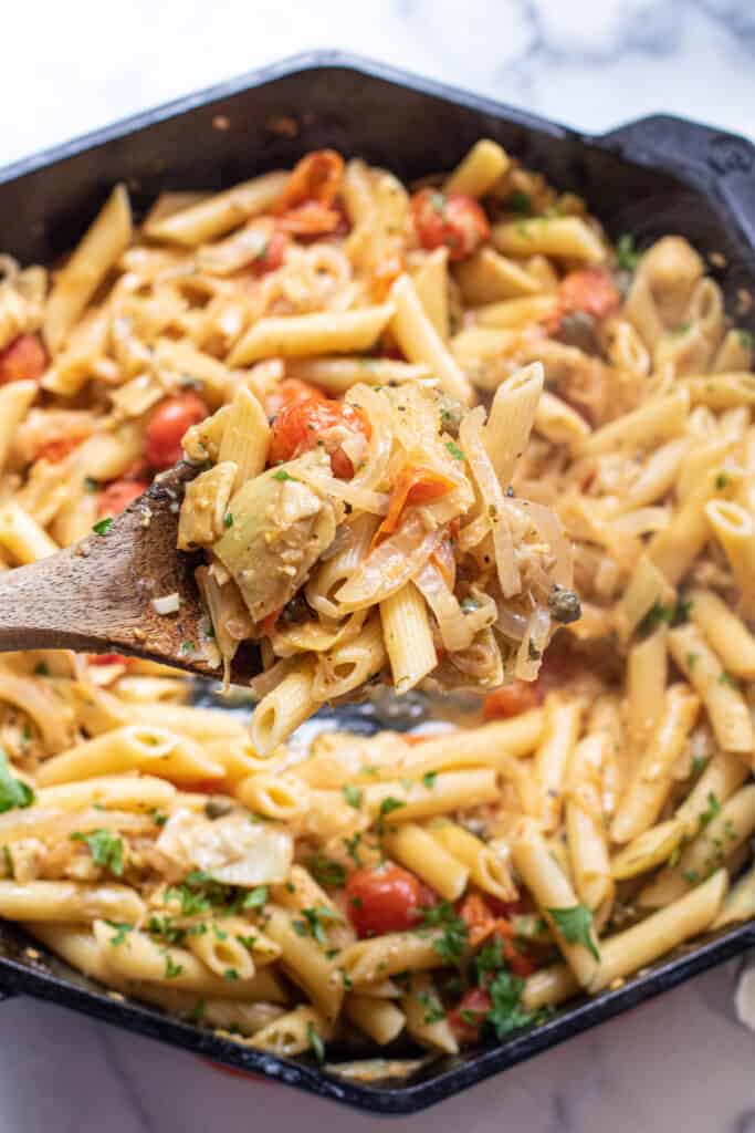 a cast iron of penne pasta with tomatoes and artichokes and parsley with a wooden spoon.