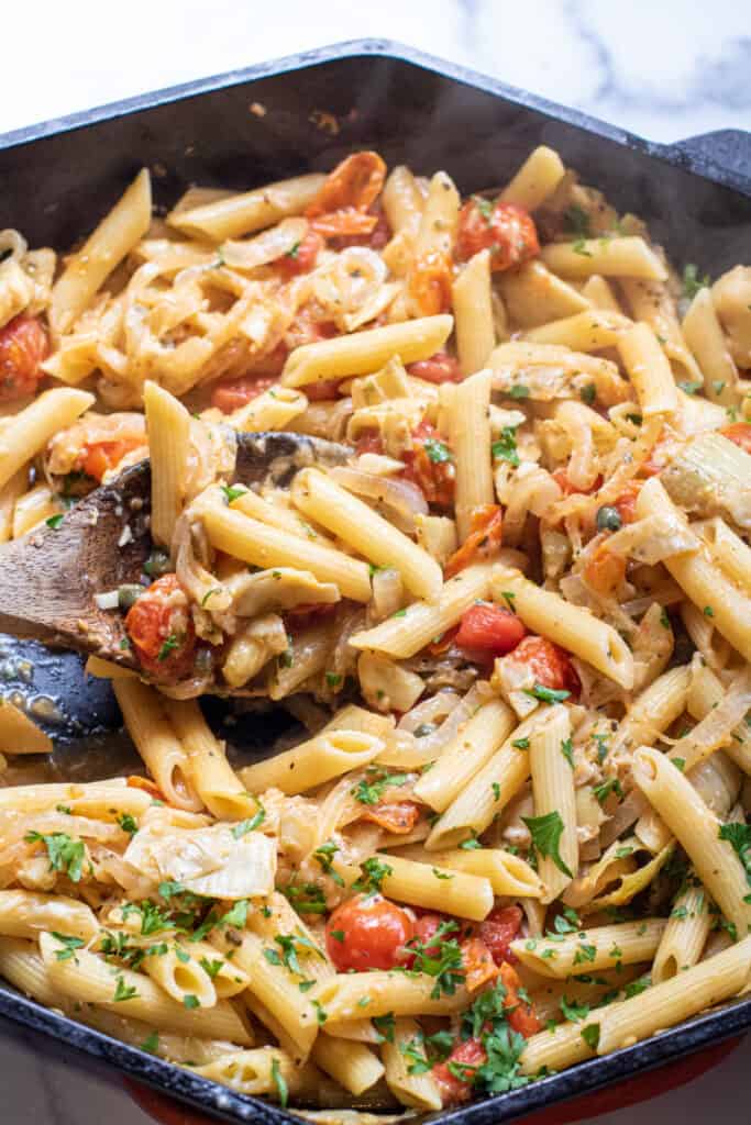 a cast iron of penne pasta with tomatoes and artichokes and parsley with a wooden spoon.