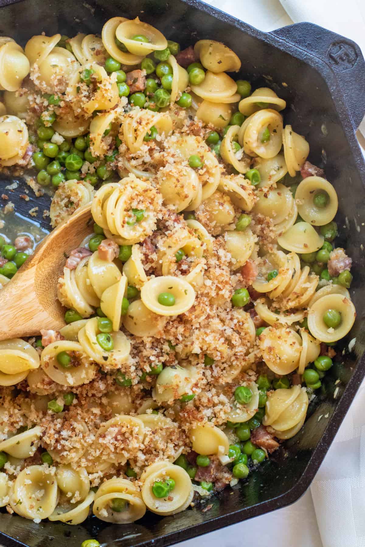 a cast iron with a wooden spoon scooping pasta with peas and pancetta.