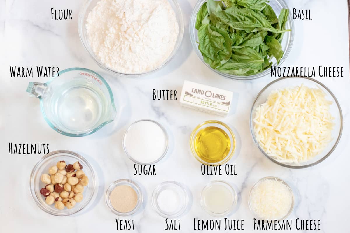 flour, basil, cheeses, sugar, butter, water, hazelnuts, oil, sugar, yeast, lemon juice all in little bowls on a counter.