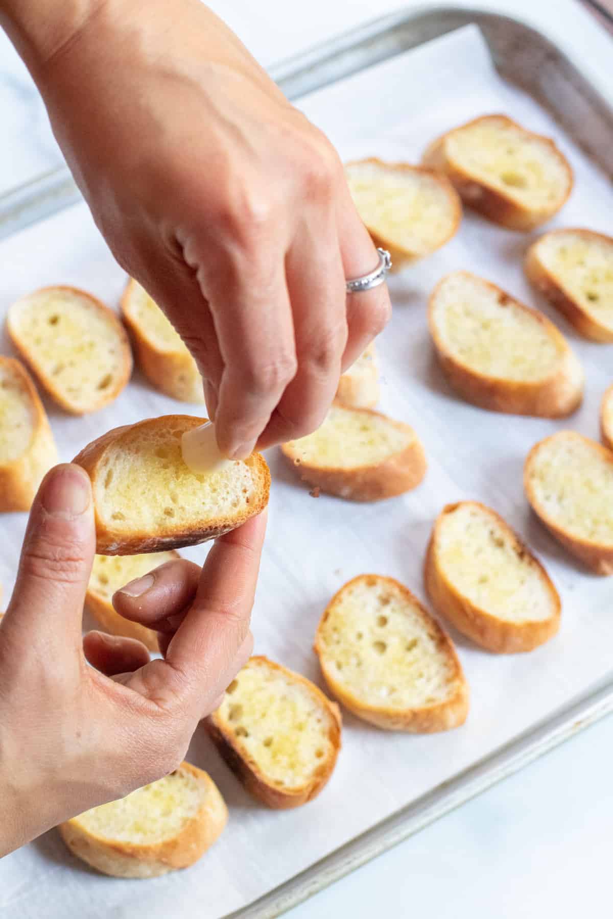 two hands holding a toasted baguette slice with fresh garlic clove being scraped on.