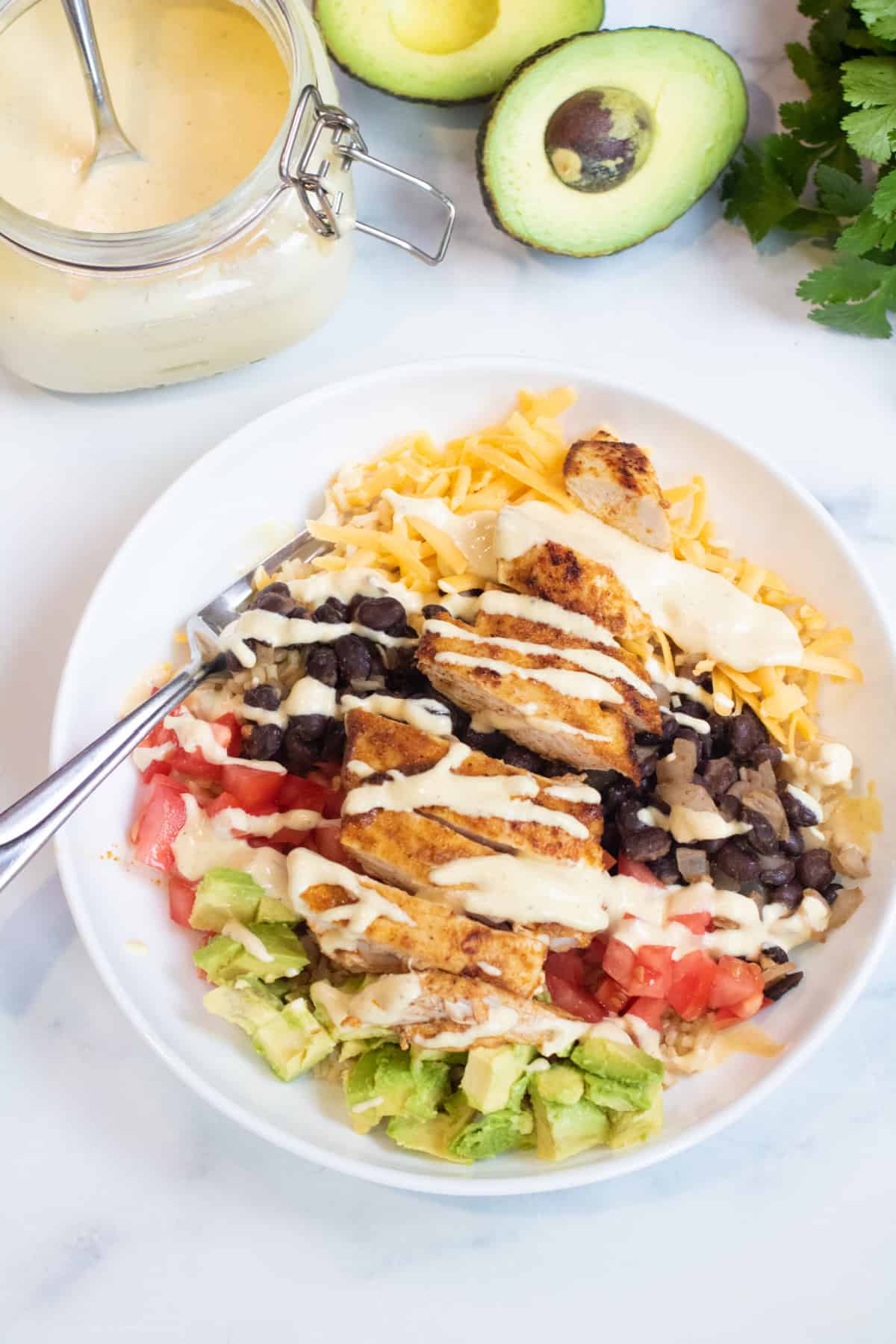 an overhead of a bowl of rice, cheese, avocado, tomatoes, black beans, chicken, and sauce.