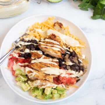 an overhead of a bowl of rice, cheese, avocado, tomatoes, black beans, chicken, and sauce.