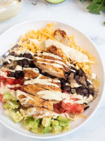 an overhead of a bowl of rice, cheese, avocado, tomatoes, black beans, chicken, and sauce.