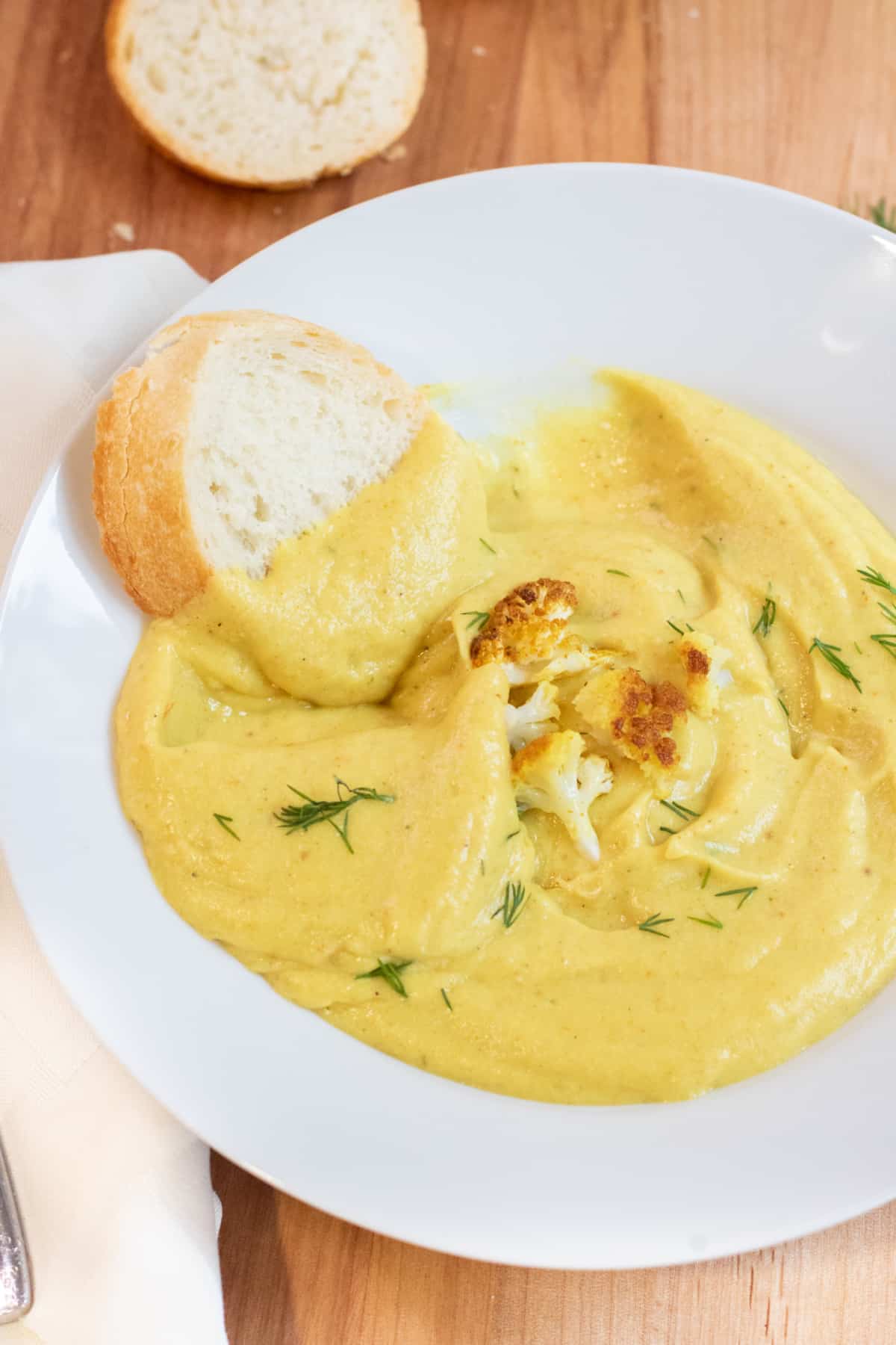 a bowl of creamy yellow soup with dill and cauliflower with a piece of baguette in it.