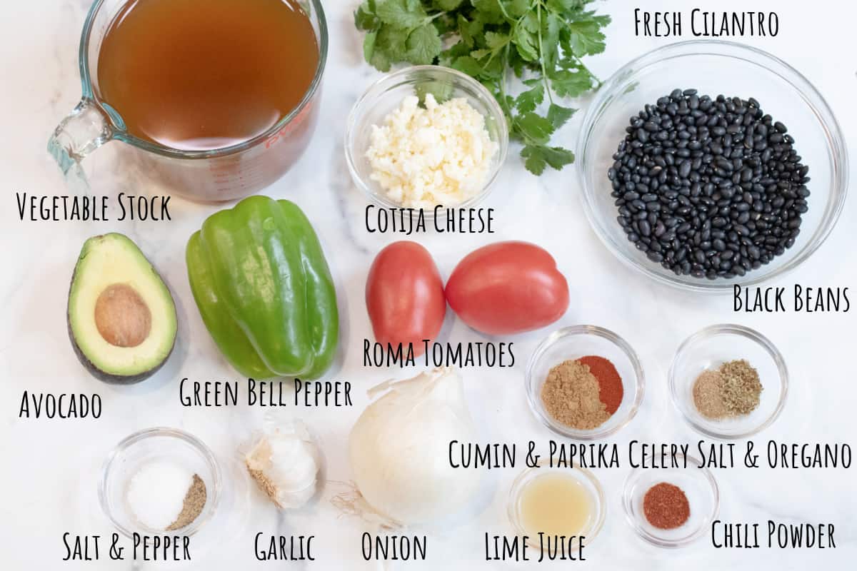 vegetable stock, dried black bens, avocado, bell pepper, tomatoes, cheese, cilantro, spices, onion and garlic on a counter all labeled.