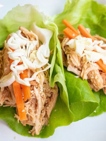 a plate of lettuce wraps with shredded chicken topped with carrots and cabbage.