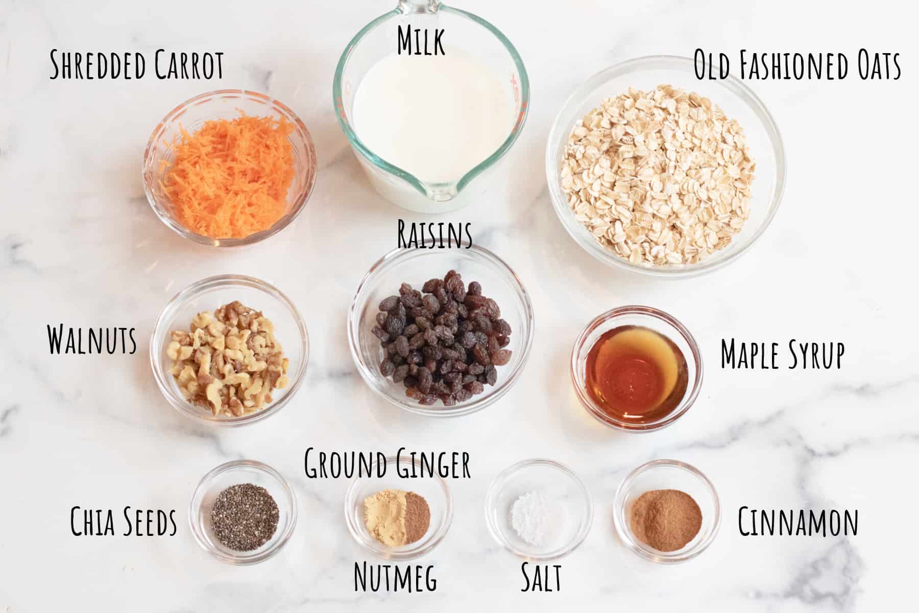 a bowl of shredded carrots, milk, oats, nuts, raisins, maple syrup, spices, on a counter.