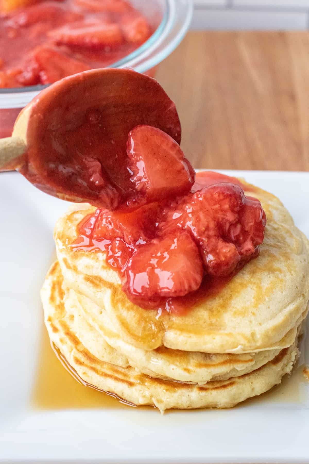 a wooden spoon scooping strawberries on a stack of pancakes.