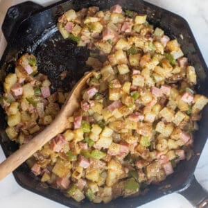 an overhead of a cast iron with a wooden spoon with small diced potato hash with ham.