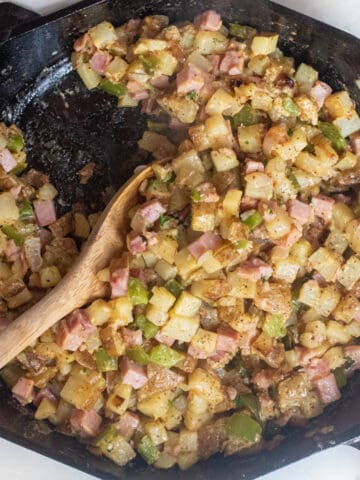 an overhead of a cast iron with a wooden spoon with small diced potato hash with ham.