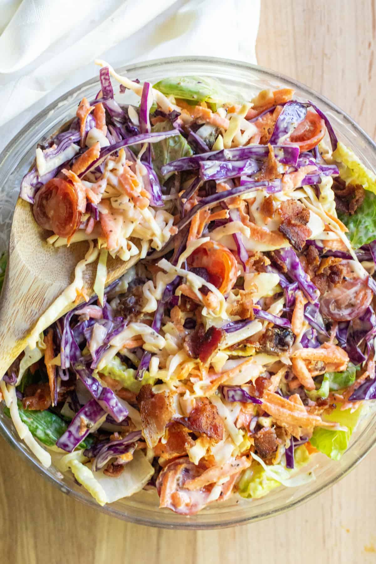 a bowl of coleslaw with bacon tomatoes and some lettuce and a wooden spoon.