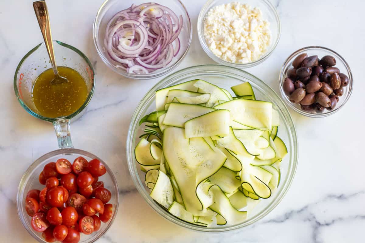 sliced zucchini in a bowl, tomatoes, onions, fetas, olive and dressing in bowls.
