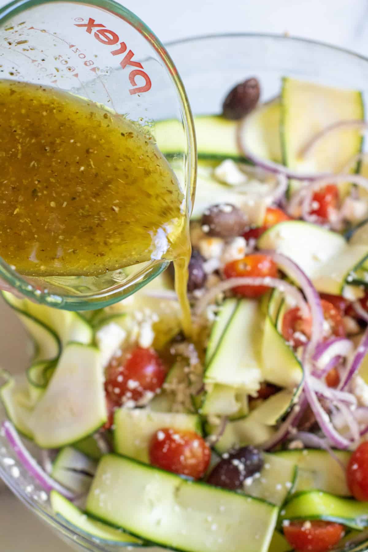 dressing being poured over a bowl of zucchinis and tomatoes and olives and onions.