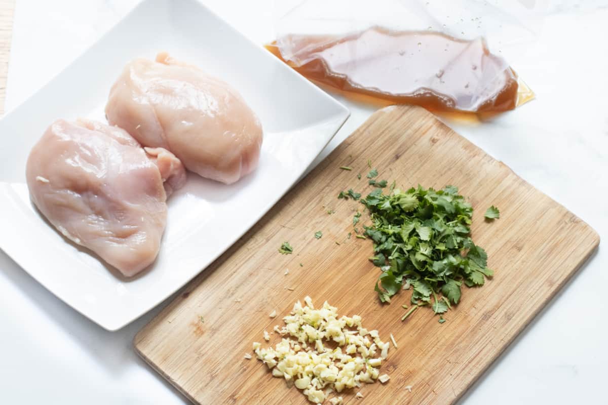 a plate of raw chicken and a cutting board with cilantro and garlic and a bag with liquid.