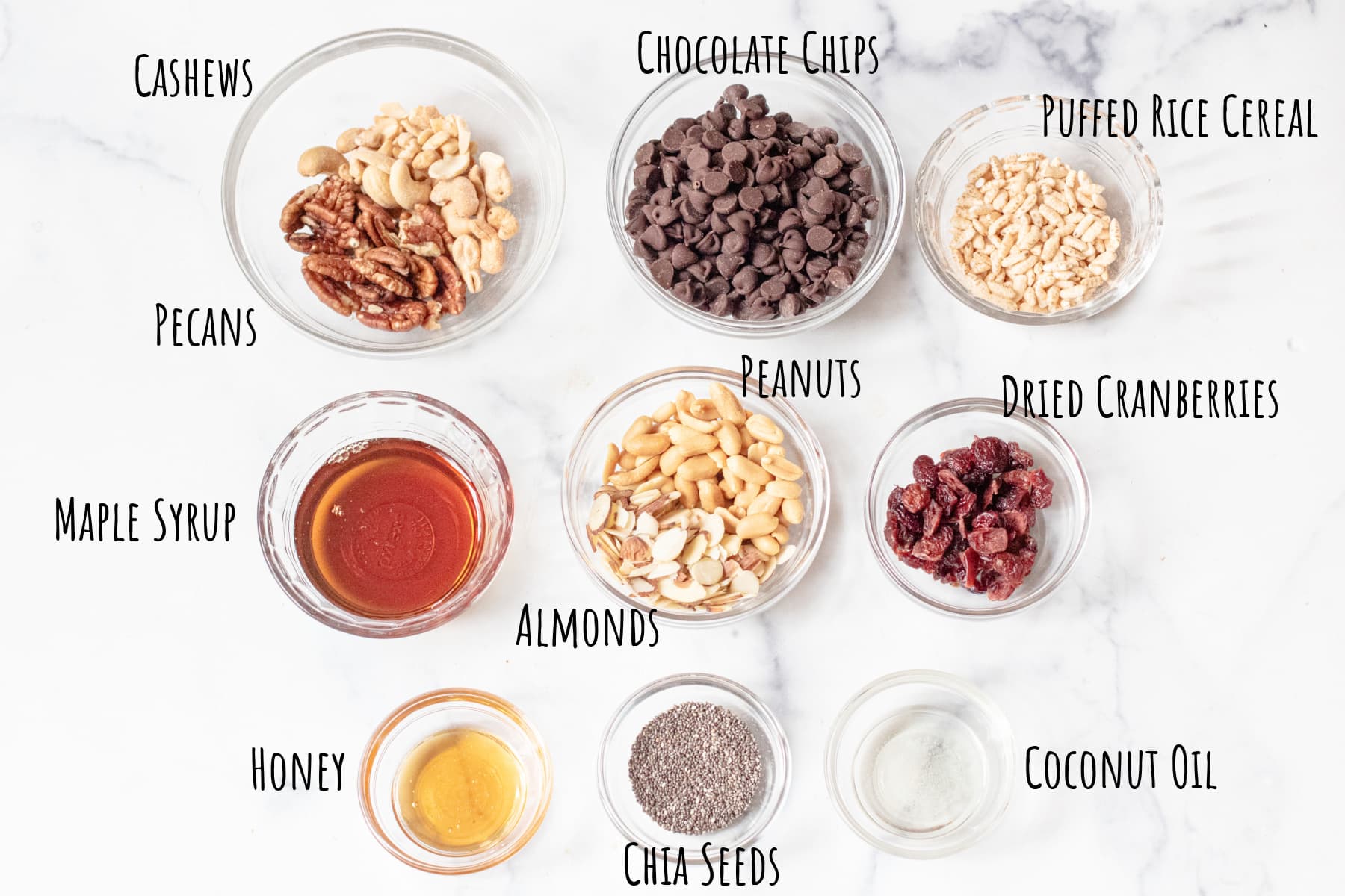 bowls of nuts, chocolate chips. rice cereal, maple syrup, cranberries, honey, chia seeds, and coconut oil.